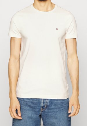 Homme portant un t-shirt uni écru à manches courtes avec un petit logo Tommy Hilfiger sur le côté gauche de la poitrine, associé à un jean bleu.