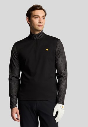 Zwarte rits-trui met patroon op de mouwen, voorzien van een gouden logo op de borst en een textuurontwerp op de armen. Witte golfhandschoen aan de linkerhand.