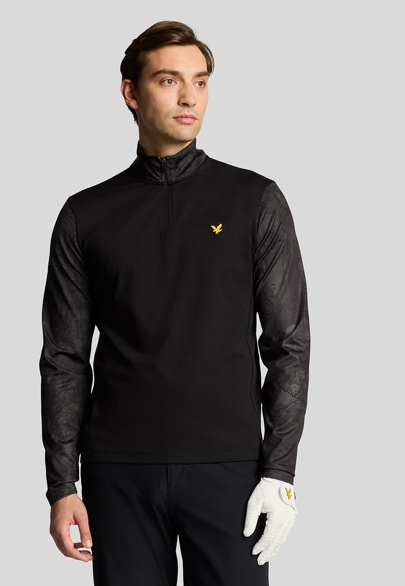 Sort zip-up pullover med mønstrede ærmer, prydet med et guld-logo på brystet og et struktureret design på armene. Hvid golfske på venstre hånd.