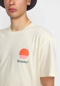 Camiseta de manga corta color crema con un gráfico circular en naranja y rosa que lleva la palabra "Revolución" en verde oscuro debajo.