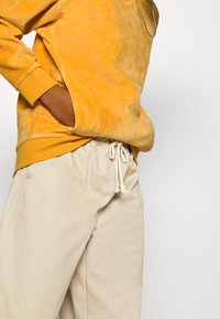Felpa in velluto giallo con polsini a coste, abbinata a pantaloni beige chiaro con cordino in vita. Nota la tasca sui fianchi e il tessuto testurizzato.