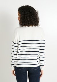 Mujer con cabello rizado que lleva un suéter blanco con rayas horizontales en azul marino y jeans oscuros, de espaldas frente a un fondo blanco liso.