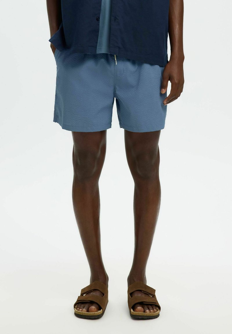 Mies, jolla on siniset, kuvioidut shortsit, tummansininen paita ja ruskeat kaksirihmasandaalit, seisoo yksinkertaisen valkoisen taustan edessä.