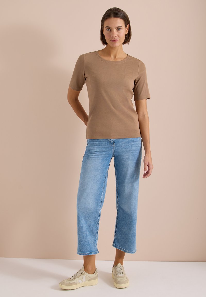 Femme debout portant une chemise marron à manches courtes, un jean court bleu clair et des baskets beiges sur un fond beige.