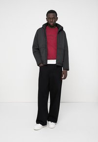 Casaco preto com capuz e acabamento texturizado, usado sobre um sweatshirt vermelho e uma camisa branca, combinado com calças largas pretas e ténis brancos.