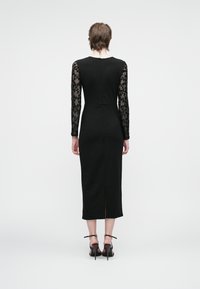 Nais, kes kannab musta midi kleiti, millel on läbipaistvad pikad varrukad, mis on kaunistatud mustade lilledega tikandiga, seisab seljaga poole ja kannab musti kõrge kontsaga sandaale.