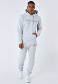 Lichtgrijze hoodie met trekkoorden, voorzien van een voorzak en logo. Gecombineerd met bijpassende taps toelopende joggingbroeken en witte sneakers.