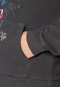 Tummanharmaa huppari, jossa on etutasku ja ribattut hihansuut. Eristävä graafinen suunnittelu, jossa on sinisiä ja vaaleanpunaisia yksityiskohtia, korostaen leikkisiä kuvioita.