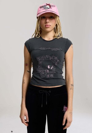 Młoda osoba z warkoczami, ubrana w różową czapkę z czaszką oraz czarny dopasowany t-shirt z haftem czaszki i kwiatów.