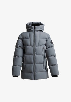 Tiener draagt een grijze gevoerde winterjas met capuchon, zwarte broek en zwarte sneakers, staand tegen een effen lichte achtergrond.