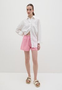 Camisa blanca de botones con un bolsillo en el pecho, combinada con pantalones cortos a cuadros rosa y sandalias de plataforma beige con detalles en las correas.