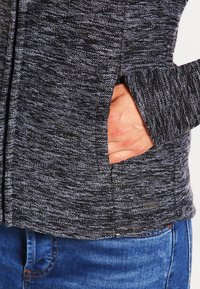 Main insérée dans la poche d'une veste texturée gris foncé au-dessus d'un jean en denim bleu.
