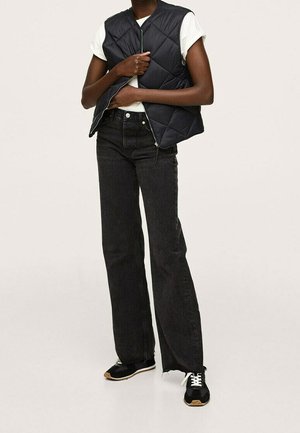 Persoon die een wit shirt met korte mouwen draagt, een zwart doorgestikt vest, zwarte jeans met brede pijpen en zwarte sneakers met witte veters.