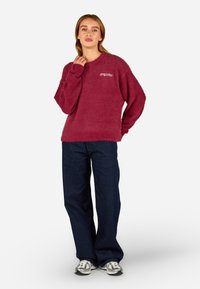 Sweater de malha felpuda em burgundy com um corte descontraído, mangas longas e um pequeno logotipo no peito, combinado com jeans de perna larga de um azul escuro e ténis prateados.