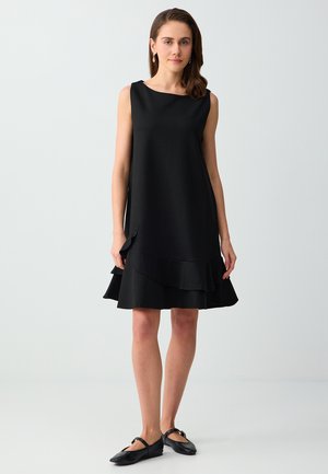 Donna in piedi che indossa un vestito nero senza maniche con orlo a balze stratificate e ballerine nere Mary Jane su uno sfondo chiaro uniforme.