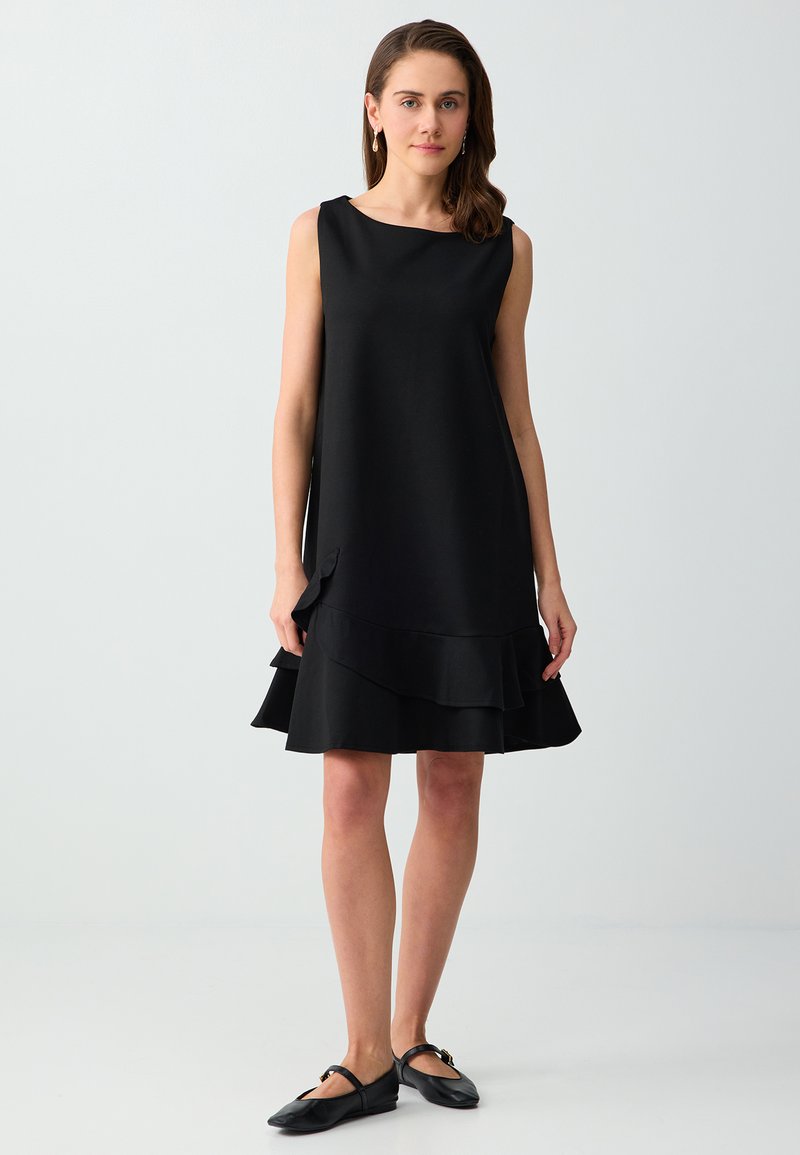 Femme debout portant une robe noire sans manches avec un ourlet à volants superposés et des ballerines Mary Jane noires sur un fond clair uni.