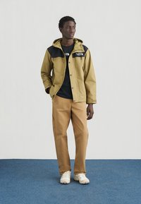 Κίτρινο και μαύρο μπουφάν με κουκούλα, κουμπιά και λογότυπο. Μαύρο πουκάμισο από κάτω, μπεζ παντελόνι και άσπρα παπούτσια σε μπλε χαλί μπροστά σε ουδέτερο φόντο.