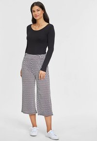 Schwarzes langärmeliges Oberteil kombiniert mit gemusterten, gekürzten Hosen mit Zickzackmuster in Weiß, Blau und Rot. Weiße Sneaker vervollständigen das Outfit.