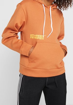 Osoba ubrana w pomarańczową bluzę z kapturem z napisem „THE THREE STRIPES” oraz czarne spodnie z białymi bocznymi pasami, ręce w kieszeni bluzy.