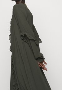 Larga y fluida vestido de gasa de un verde oliva oscuro, con bordes rizados en las mangas y a lo largo del costado. Corte suelto y escote redondo.