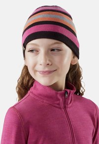 Ragazza che indossa una giacca a zip viola e un berretto a righe nei colori rosa, arancione, nero e grigio, guarda leggermente verso destra con un sorriso.