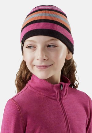 Chica con una chaqueta morada con cremallera y un gorro a rayas en rosa, naranja, negro y gris, mirando ligeramente hacia la derecha con una sonrisa.
