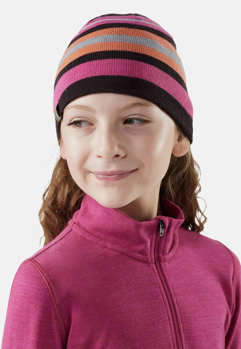 Ragazza che indossa una giacca a zip viola e un berretto a righe nei colori rosa, arancione, nero e grigio, guarda leggermente verso destra con un sorriso.