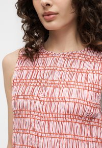 Femme portant un haut rose sans manches avec des plis verticaux texturés et des coutures horizontales orange, cheveux bruns bouclés visibles aux épaules.
