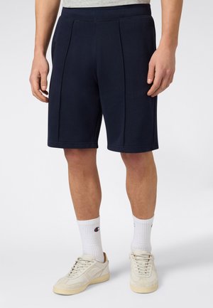 Uomo con pantaloncini blu navy fino al ginocchio, calzini bianchi alla caviglia e sneakers color avorio, in piedi davanti a uno sfondo uniforme.