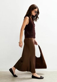 Woman walking in brown sleeveless cardigan and long skirt, black flats, sunglasses, and carrying large brown handbag.