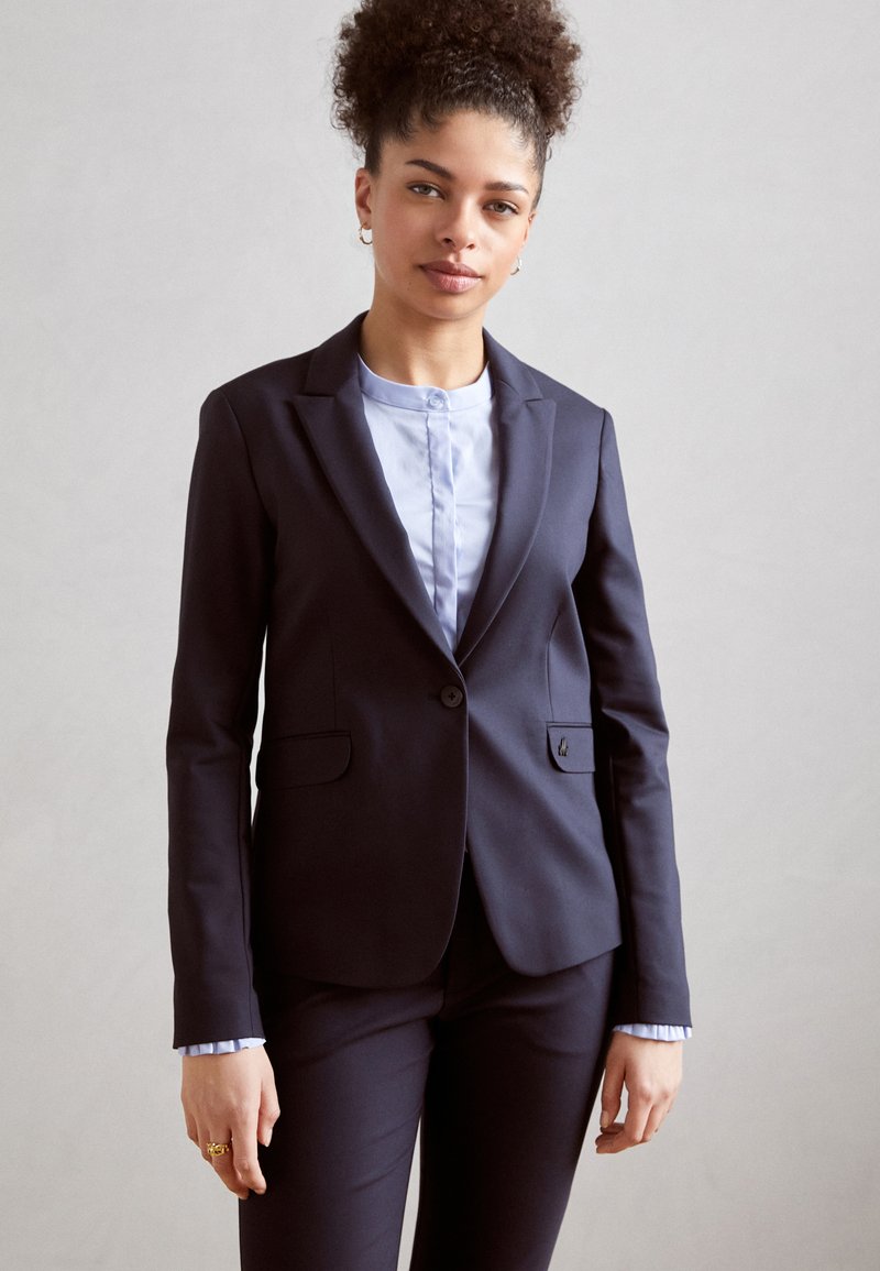 Námořnický saka s jedním knoflíkem, dvěma předními kapsami a elegantními klopami, kombinované se světle modrou blůzou a odpovídajícími kalhotami.