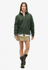 Sudadera de forro polar verde oscuro con cremallera de cuarto, con puños acanalados, combinada con una camisa blanca y una falda de cargo color caqui. Botas de cuero negras hasta la rodilla.