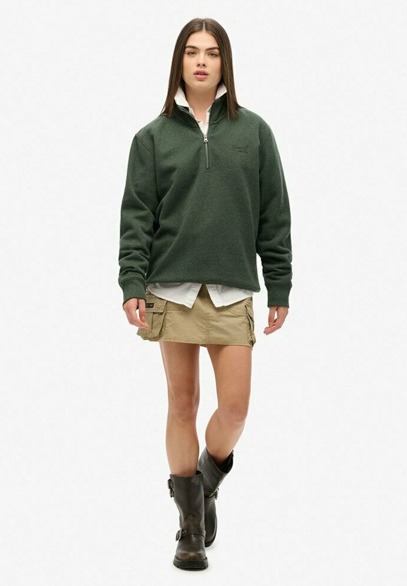 Sudadera de forro polar verde oscuro con cremallera de cuarto, con puños acanalados, combinada con una camisa blanca y una falda de cargo color caqui. Botas de cuero negras hasta la rodilla.