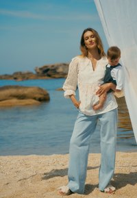 Witte blouse met oogjes en een diepe V-hals, gecombineerd met lichtblauwe wijde jeans en beige sandalen, terwijl ze een kind bij de waterkant vasthoudt.