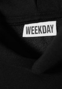 Schwarzes Stoffetikett mit dem Aufdruck "WEEKDAY" in fettgedruckten weißen Buchstaben. Weiche Textur, mit einem leicht abgenutzten Rand am Kragen.
