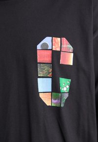 Musta t-paita, jossa on suuri graafinen kirjain "C", joka on koostettu värikkäistä, tilkkutäkki-tyylisistä kuvista eri muodoissa ja tekstuureissa.