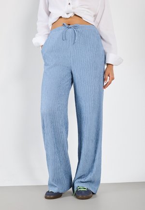 Frau trägt hellblaue, gerippte, weit geschnittene Hose mit Kordelzug in der Taille, kombiniert mit marineblauen Sneakers und einem weißen, geknoteten Hemd.