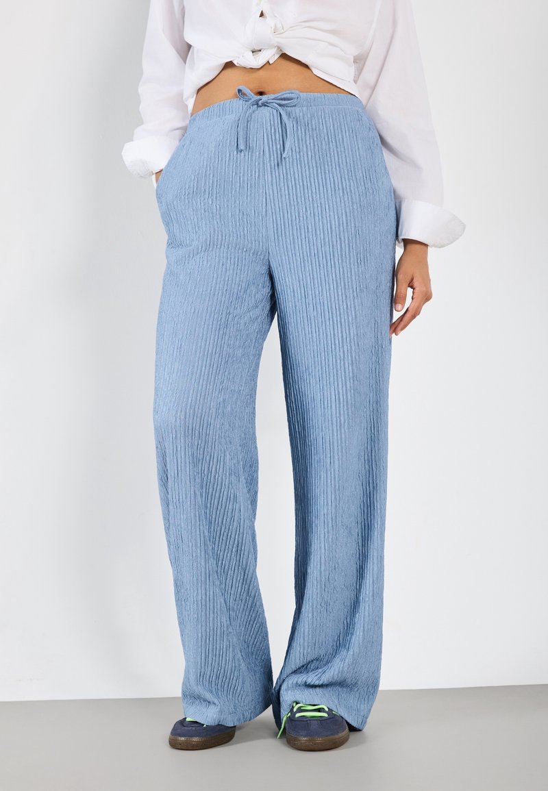 Femme portant un pantalon large côtelé bleu clair avec taille cordon, associé à des baskets bleu marine et une chemise blanche nouée.