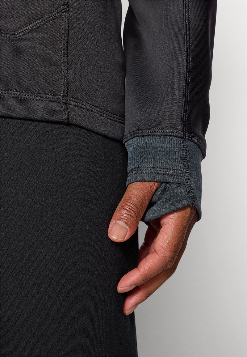 Maglia sportiva a maniche lunghe nera, con polsino testurizzato in grigio scuro. Primo piano di una mano che afferra il tessuto, evidenziando le cuciture.