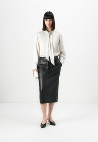 Femme avec des cheveux raides et foncés, portant un blouse en satin blanc et une jupe crayon en cuir noir, tenant un petit sac à main blanc, se tenant devant un fond blanc.