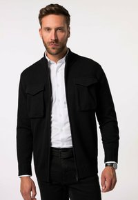 Schwarze Strickjacke mit Reißverschluss, zwei Brusttaschen, gerippten Bündchen und einem Kragen, getragen über einem weißen Hemd mit Knöpfen und schwarzen Hosen.