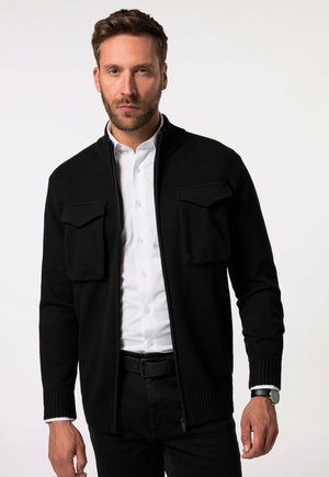 Zwarte gebreide cardigan met een rits, twee borstzakken, geribde manchetten en een kraag, gedragen over een witte overhemd en zwarte broek.