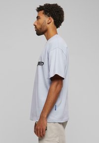 Lichtgrijze katoenen t-shirt met korte mouwen, ronde halslijn en een zwart logo op de borst; heeft een blauw label aan de onderkant.