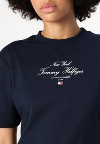 Tamno plava pamučna majica s natpisom "New York Tommy Hilfiger" u bijelom rukopisnom fontu, s logom i detaljem "All American Classics" ispod.