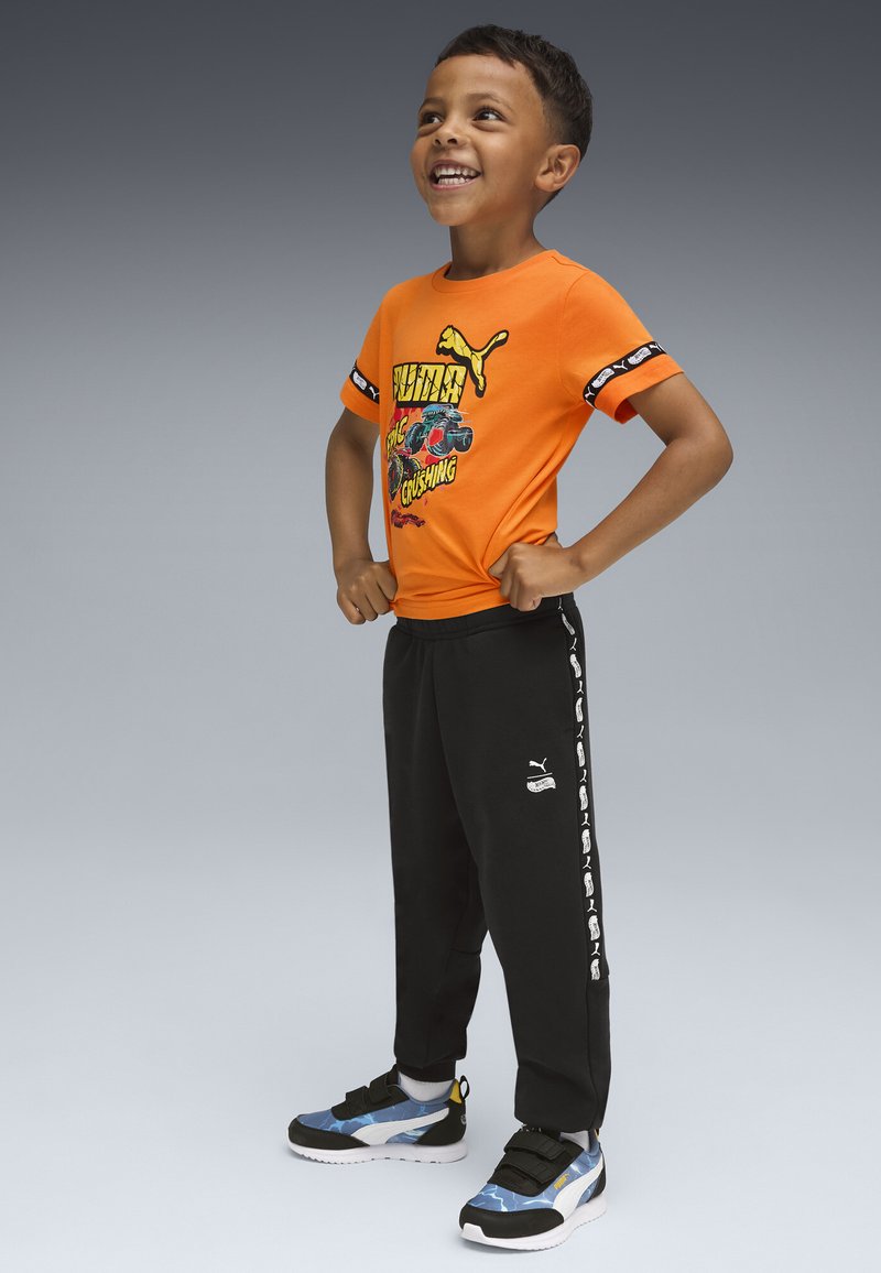 Junger Junge mit einem Lächeln und Händen in den Hüften, trägt ein orangefarbenes Grafik-T-Shirt, schwarze Sweatpants mit seitlichen Logos und blaue Turnschuhe vor einem grauen Hintergrund.