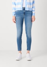 Jean skinny bleu en denim, avec une taille mi-haute, longueur cheville, et une texture légèrement décolorée, associés à des baskets blanches.