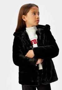 Sort faux fur jakke med hætte, der har en knaplukning og en blød overflade. Bæres over en hvid sweatshirt med et rødt grafisk design.