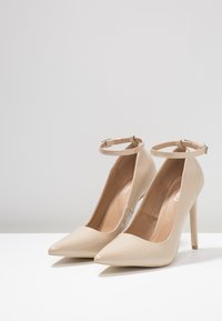 Beige lackläderpumps med hög klack, spetsiga tår och ankelremmar. Slät yta, smala klackar och minimalistiska designdetaljer.