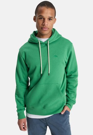 Junger Mann trägt einen grünen Kapuzenpullover mit weißen Kordeln und blaue Hosen, steht mit einer Hand in der Tasche vor einem schlichten Hintergrund.