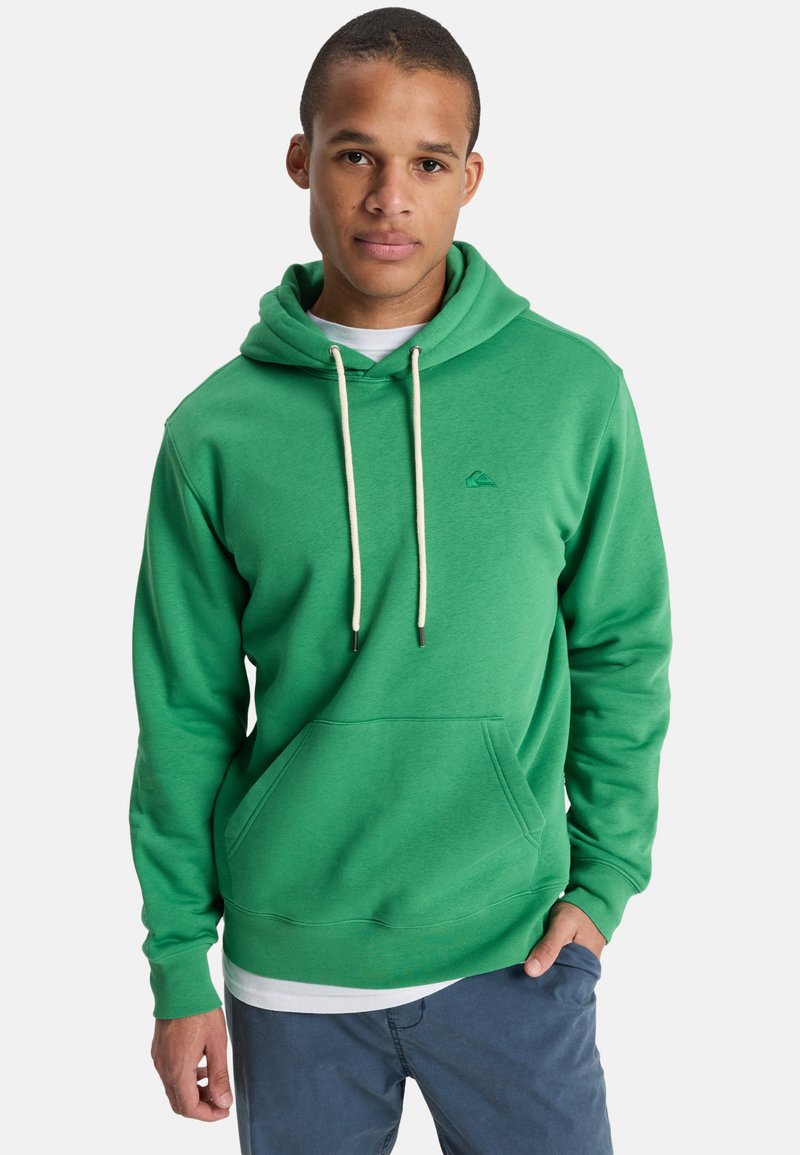 Giovane uomo con indosso una felpa verde con coulisse bianche e pantaloni blu, in piedi con una mano in tasca contro uno sfondo semplice.